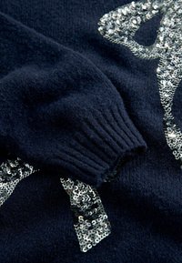 Mørkeblå strikket sweater med ribbet manchet, prydet med et mønster af sølvfarvede pailletter arrangeret i et design på stoffet.