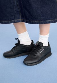 Baskets en cuir noir avec une finition texturée, dotées d'un design à lacets et d'un col rembourré. Portées avec des chaussettes blanches et une jupe en jean.