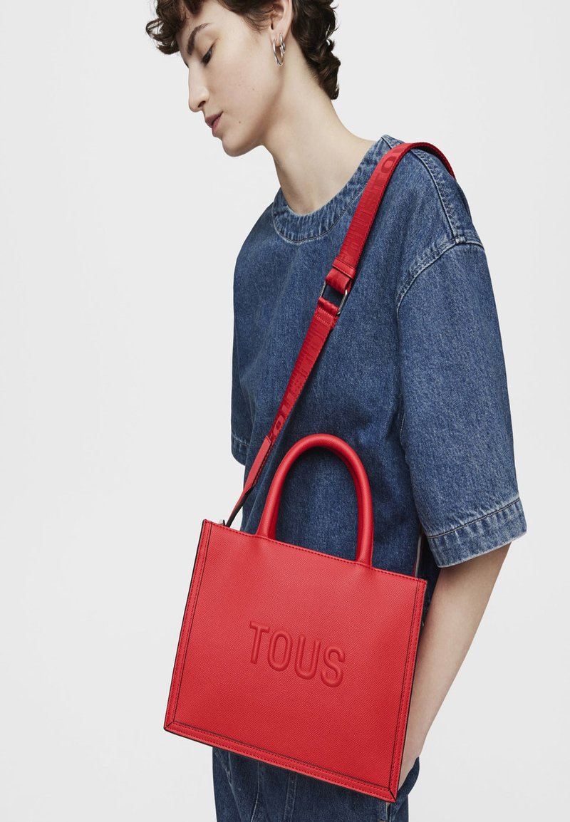 Bolso de mano de cuero rojo con el logo "TOUS" en relieve, de forma rectangular, con dos asas superiores y una correa ajustable. Acabado texturizado y líneas limpias.