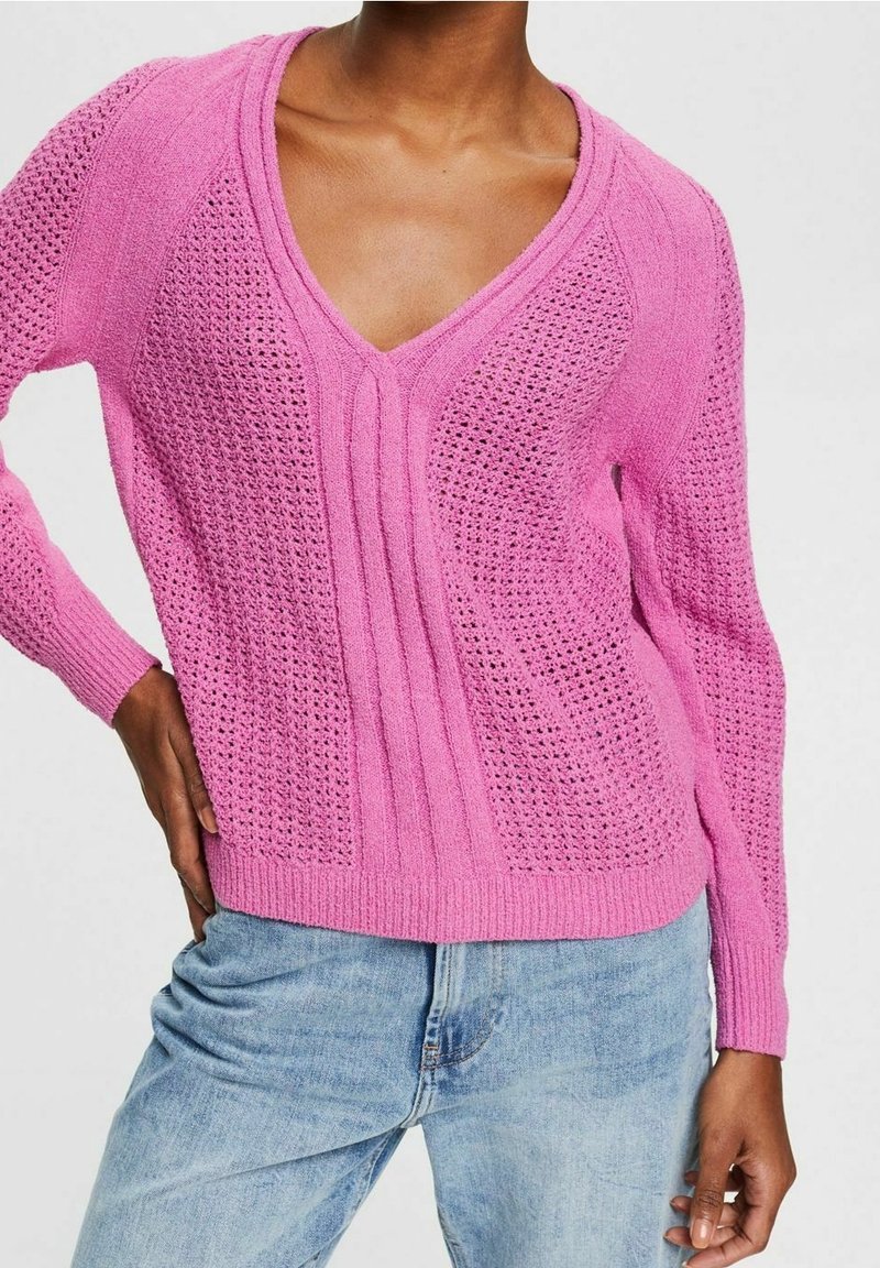Femme portant un pull en tricot rose vif à col en V avec des motifs texturés, associé à un jean en denim bleu clair, main sur la hanche.