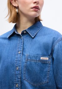 Γυναίκα με μπλε πουκάμισο τζιν με κουμπιά και τσέπη με την ετικέτα "Calvin Klein" και ασημένια σκουλαρίκια, σε απλό φόντο.