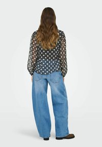Femme avec de longs cheveux ondulés portant un chemisier noir transparent à pois et un jean bleu clair à jambes larges, se tenant de dos sur un fond uni.