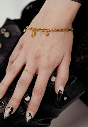 Hand with black and white abstract nail art holding a black textured purse, wearing a delicate gold chain bracelet with charms and a thin gold ring.