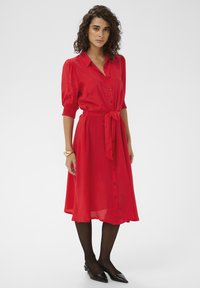 Robe rouge boutonnée avec manches courtes bouffantes et ceinture assortie. Tissu léger avec une jupe fluide, associée à des chaussures noires à bouts pointus.