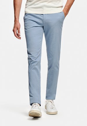Homme portant un pantalon slim bleu clair et des baskets blanches, marchant avec une main dans la poche sur un fond blanc.