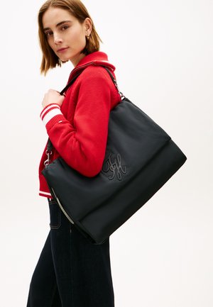 Ung kvinne iført en rød genser holder en stor svart totebag med dekorative sømmer på siden, stående mot en ensfarget bakgrunn.