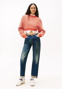 Rövidített narancssárga és fehér csíkos pulóver, magas derekú kék farmernadrág szakadt szegéllyel és fehér sportcipő kék díszítéssel.