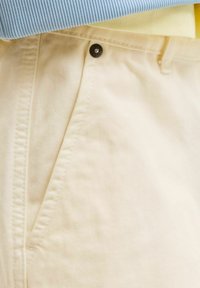 Gros plan sur un pantalon beige clair avec un bouton rivet en métal, des surpiqûres sur la poche et un tissu superposé bleu et jaune au-dessus.