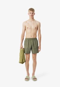 Zwemshorts in olijfgroen met een golvend patroon, elastische tailleband en zijsplitten. Houdt een lichtgroen shirt vast en draagt lichtgroene slippers.