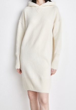 Mujer con un vestido de punto oversize color blanco hueso con capucha y puños y dobladillo acanalados, posando frente a un fondo blanco liso.