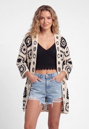 Vrouw draagt een zwart kanten crop top, lichtblauwe gescheurde denimshorts en een lange zwart-witte gehaakte cardigan met patroon.