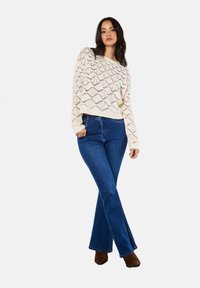Krämfärgad stickad tröja med diamantmönster, matchad med blå utställda jeans. Modellen står med händerna på höfterna, iförd bruna anklar stövlar.