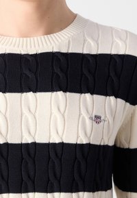 Personne portant un pull en maille torsadée crème et noir avec des rayures horizontales et un petit logo écusson brodé sur la poitrine.