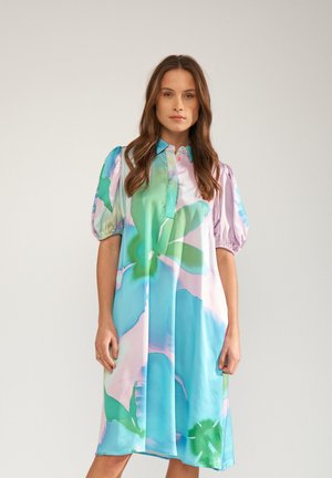 Hosszú barna hajú nő, aki egy térdig érő pasztell kék-zöld virágos ruhát visel puffos ujjakkal és gallérral, egy egyszerű háttér előtt áll.