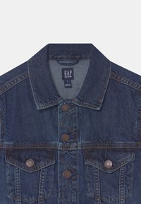 Tmavě modrá džínová bunda s límcem, která má dvě náprsní kapsy, hnědé kovové knoflíky a viditelné švy. Štítek nese nápis "GAP DENIM."