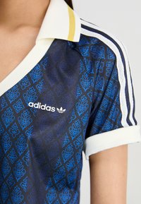 Mörkblå kortärmad skjorta med blommönster i blått, vita accenter och en vit krage med ett guldstripe; har Adidas-logotyp.