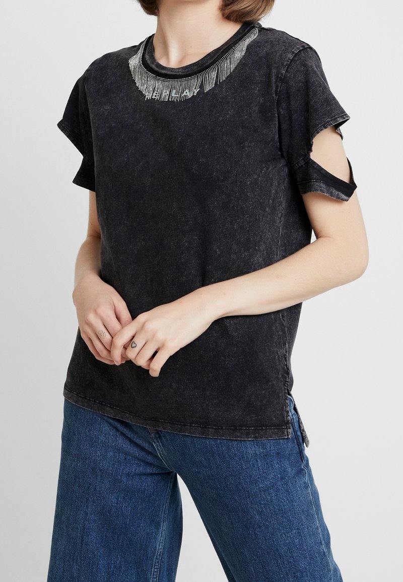 Camiseta negra de manga corta con un aspecto desgastado, que presenta un detalle de flecos metálicos alrededor del escote y un diseño de hombros descubiertos.