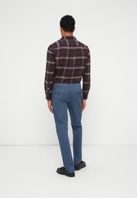 Camicia a maniche lunghe a quadri nei toni del rosso scuro e blu, abbinata a pantaloni blu. L'outfit presenta una vestibilità su misura e una texture liscia.