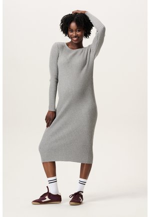 Glunderende vrouw staat met één hand op haar hoofd, gekleed in een lange grijze geribbelde jurk, gestreepte witte sokken en bordeauxrode sneakers.