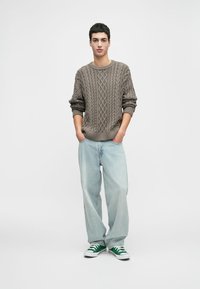 Strickpullover in Taupe mit Kabelmuster, kombiniert mit hellblauen, lässig geschnittenen Jeans und grün-weißen Sneakern. Das Model steht mit den Händen in den Taschen.