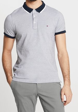 Mann trägt ein hellgraues Poloshirt mit kurzen Ärmeln, dunklem Kragen und Bündchen, kombiniert mit hellgrauen Hosen, steht mit entspannten Händen.