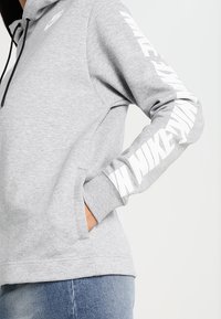Šedá mikina s kapucí a černým stahovacím lurexem. Na pravém rukávu vertikálně vytištěný bílý text "NIKE". Měkká fleece textura.