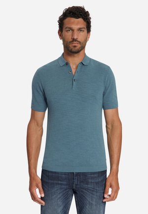 Man met een blauw poloshirt met korte mouwen en een donkerblauwe spijkerbroek, staand voor een effen witte achtergrond.