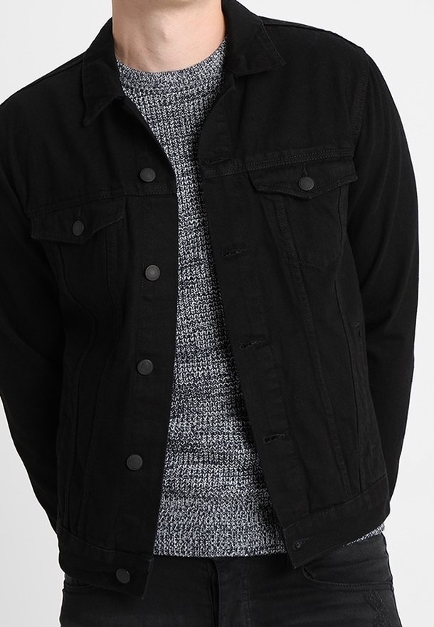 Svart denimjacka med framknappar och två bröstfickor, bärs över en strukturerad grå stickad tröja. Enkel design, inga utsmyckningar.