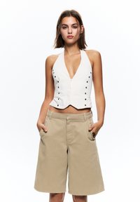 Fehér, nyakpántos crop top elülső patent gombokkal; bézs, térd térd alá érő rövidnadrággal párosítva, amelyek oldalsó zsebekkel és elülső cipzárral rendelkeznek.