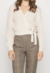 Slonovinový pletený svetr s hlubokým výstřihem do V, dlouhými rukávy a detailem vázání v pase, doplněný o hnědé kárované kalhoty s vysokým pasem.