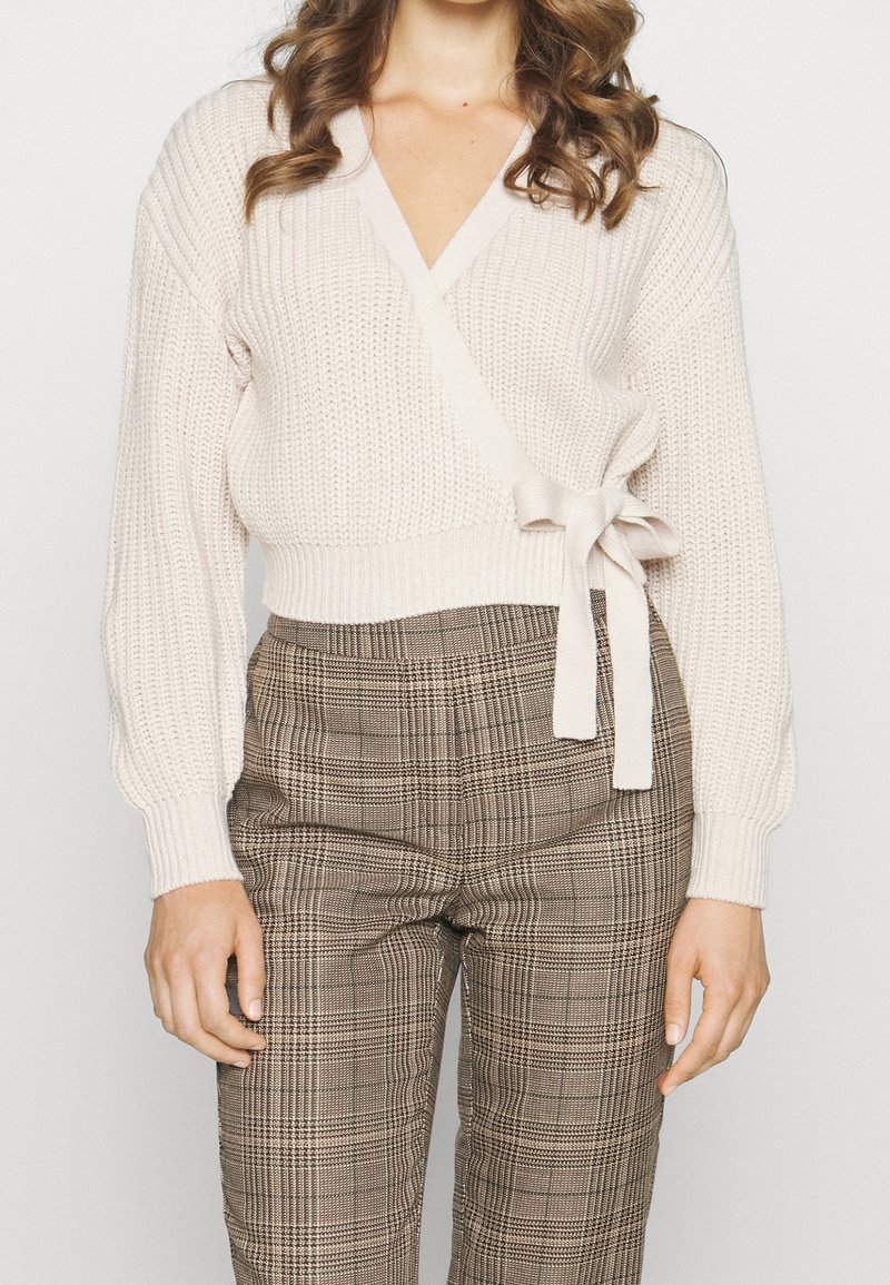 Slonovinový pletený svetr s hlubokým výstřihem do V, dlouhými rukávy a detailem vázání v pase, doplněný o hnědé kárované kalhoty s vysokým pasem.