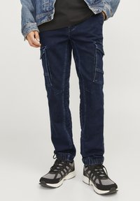 Pantalones cargo azul marino con puños elásticos, que presentan múltiples bolsillos, detalles de costura y un ajuste casual. Llevados con zapatillas deportivas negras y grises.