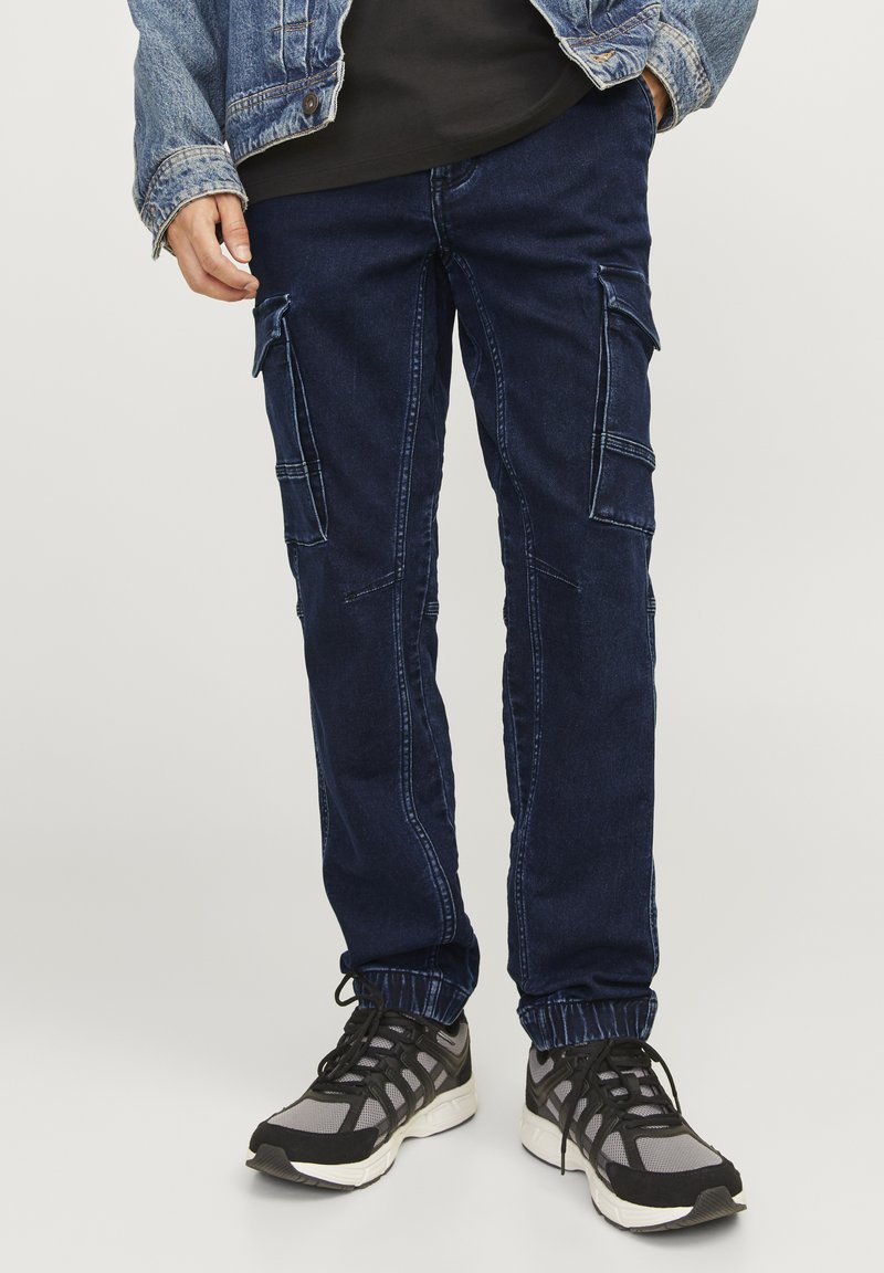 Pantalones cargo azul marino con puños elásticos, que presentan múltiples bolsillos, detalles de costura y un ajuste casual. Llevados con zapatillas deportivas negras y grises.