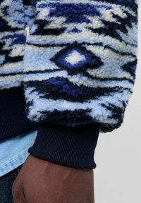 Fleece jack met geometrische patronen in donkerblauw, lichtblauw en crème. Ribgebreide marineblauwe manchetten, zachte textuur en een relaxte pasvorm.