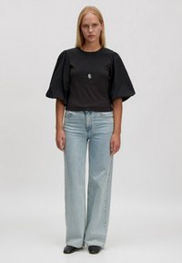 Vrouw die staat, gekleed in een zwart top met pofmouwen, lichtblauwe wijde jeans, zwarte schoenen, een zilveren hangertje als ketting en ring oorbellen.