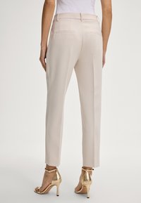 Pantaloni beige su misura con gamba dritta, due tasche posteriori, abbinati a sandali alti dorati, con una sottile cinghia alla caviglia.