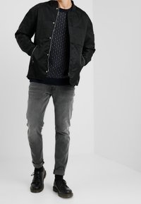 Černá prošívaná bunda s límcem na stojáka, nošená přes tmavě modrý vzorovaný svetr, doplněná šedými zúženými kalhotami a černými koženými botami na šněrování.