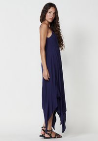 Robe sans manches bleu marine avec ourlet asymétrique. Présente de légers embellissements et un tissu doux et fluide. Associée à des sandales noires.