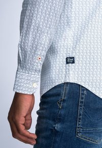 Wit overhemd met lange mouwen en een geometrisch blauw patroon. Voorzien van knopen bij de manchetten en een marineblauw label aan de zijkant. Gepaarde met donkere denim.