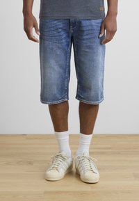 Pantalones cortos de mezclilla azul con un corte hasta la rodilla, que presentan un acabado desgastado, costuras clásicas y un diseño de pierna recta, combinados con zapatillas blancas.