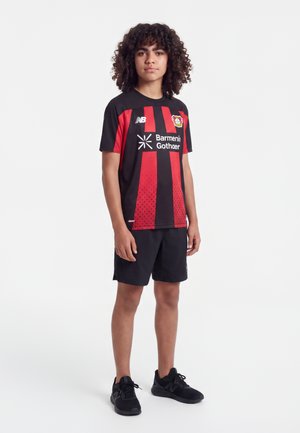 Young person with curly hair wearing a black and red Bayer Leverkusen football jersey, black shorts, and black sneakers standing on white background.