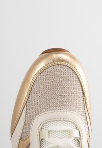 Gros plan sur la pointe d’une basket avec un liseré métallique doré, une tige en mesh beige, des lacets blancs et un panneau en tissu texturé.