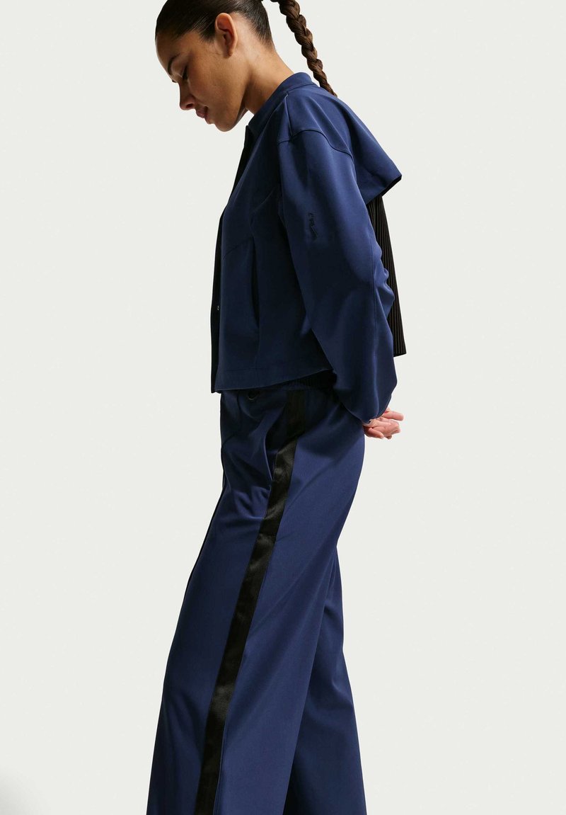 Frau mit geflochtenem Haar, die eine marineblaue Jacke und passende Hosen mit schwarzen Seitenstreifen trägt, steht mit den Händen hinter dem Rücken.