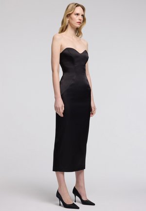 Mujer con un vestido midi negro ajustado sin tirantes y tacones altos negros, de pie contra un fondo gris claro, mirando hacia un lado.