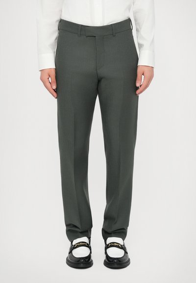 Tumši zaļi pielāgoti bikses ar gludu tekstūru, ar taisnu piegriezumu un jostas cilpām, kombinētas ar melniem mokasīniem un baltām zeķēm.