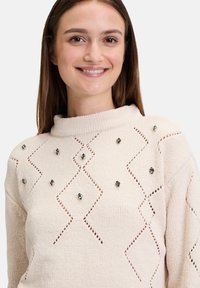 Stickad krämfärgad tröja med hög krage, prydd med öppna diamantmönster och dekorativa svarta stenar. Mjuk textur, avslappnad passform.