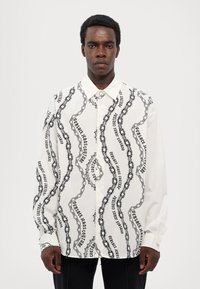 Camisa blanca de botones con patrón de cadenas negras y texto, con mangas largas y un diseño de cuello clásico. Detalle de doble puño presente.