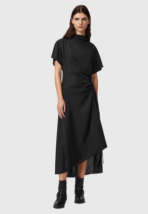 Kvinde iført en sort asymmetrisk midi-kjole med korte ærmer og sorte ankelstøvler, stående foran en ensfarvet baggrund.