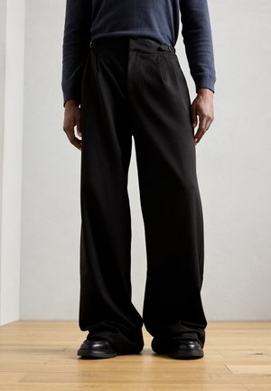 Black wide-leg trousers made of smooth fabric. Features pleats at the front and belt loops. Paired with black shoes on a wooden floor.