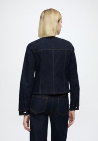 Σκούρο τζιν μπουφάν που δείχνει την πίσω όψη με εμφανείς ραφές, δύο πίσω τσέπες και μανσέτες με κουμπιά· συνδυασμένο με αντίστοιχο σκούρο τζιν παντελόνι.